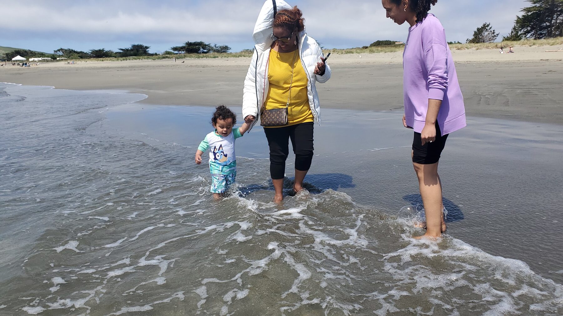 A toddler holding an adult's hand while stepping into the shallow foam of an incoming wave on a wide sandy beach