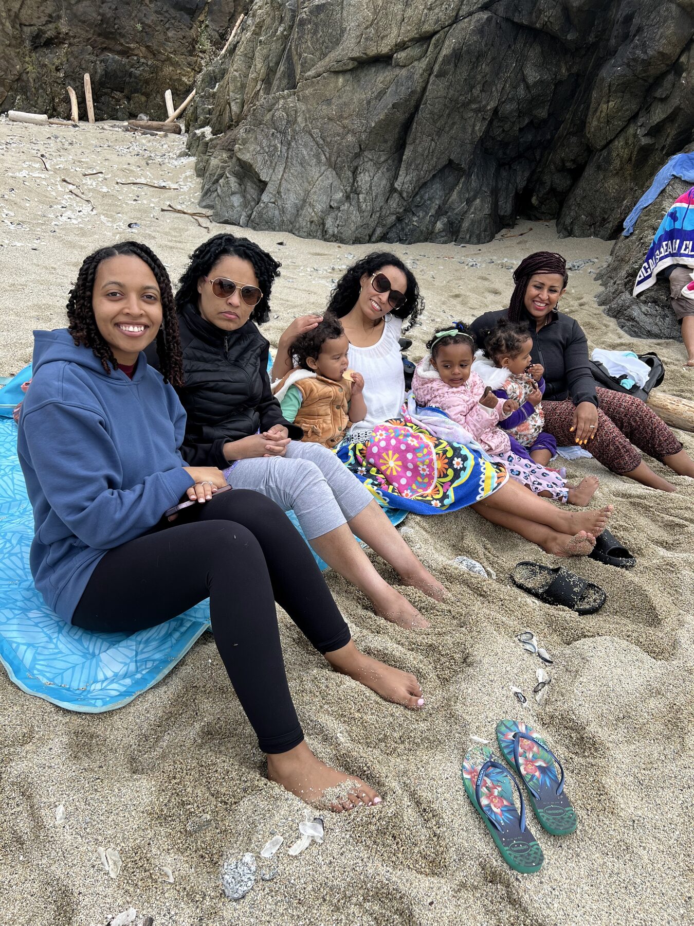 Four women sitting on beach blankets in the sand with three small children, resting beside a large rock outcrop