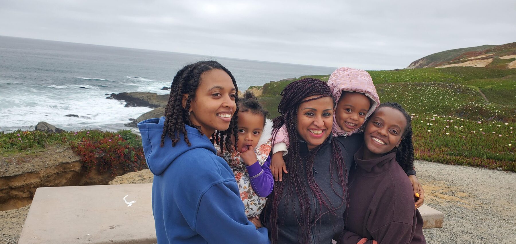Three women holding two toddlers, smiling together on a green bluff above the grey Pacific on a past family trip