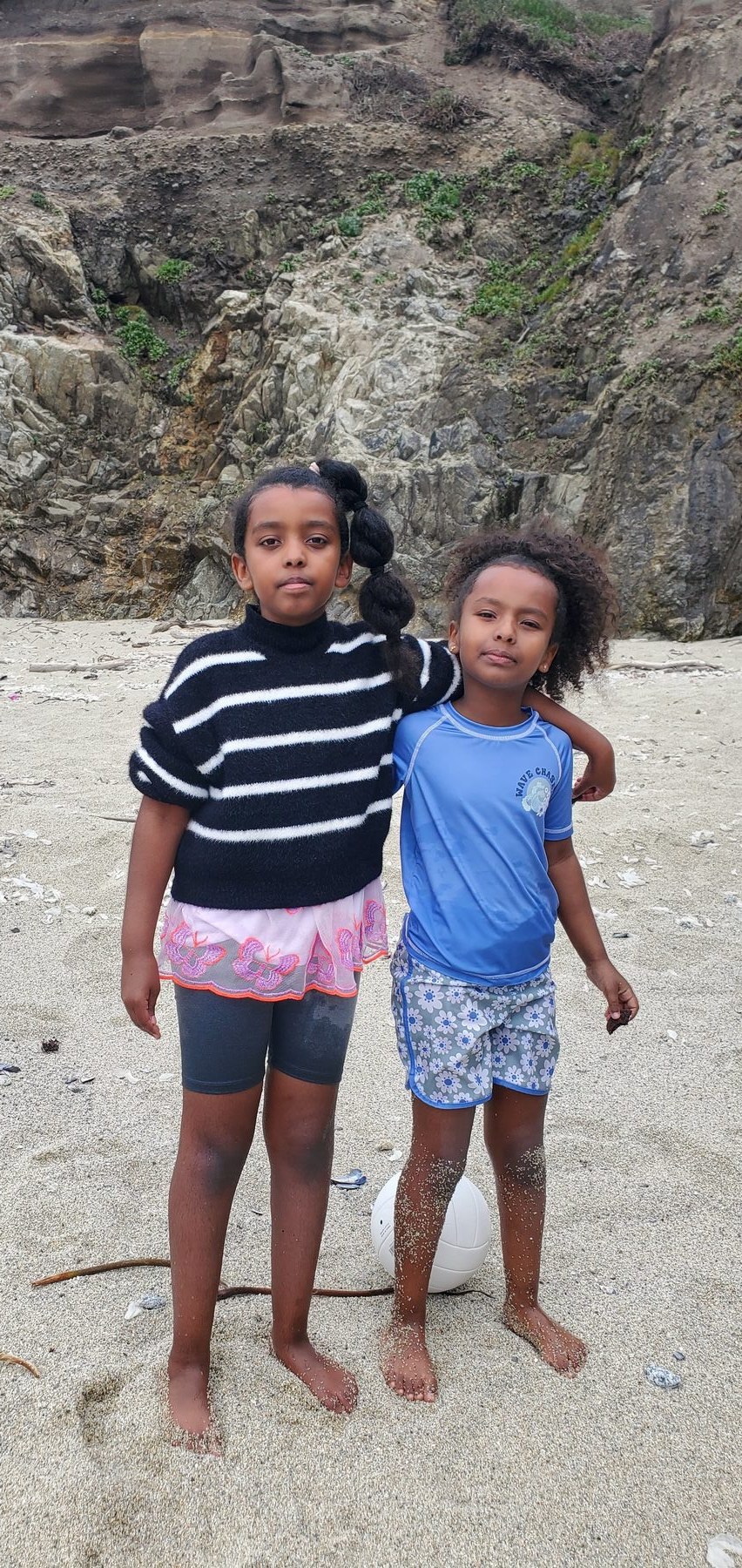 Two young girls standing barefoot on the sand beside a volleyball, looking up at the camera