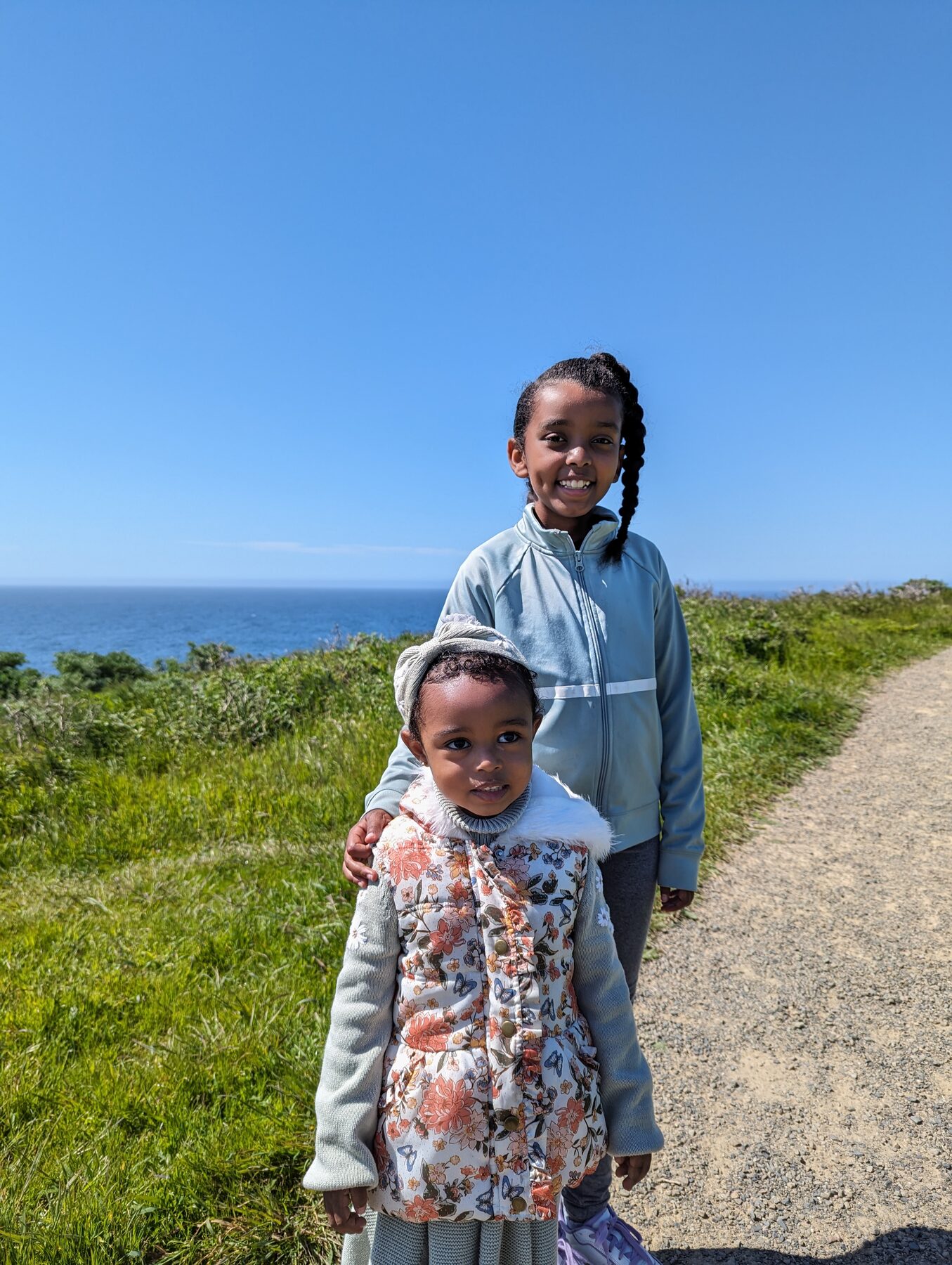 An older girl with her arm around a small toddler on a gravel coastal trail, with the deep blue ocean behind them
