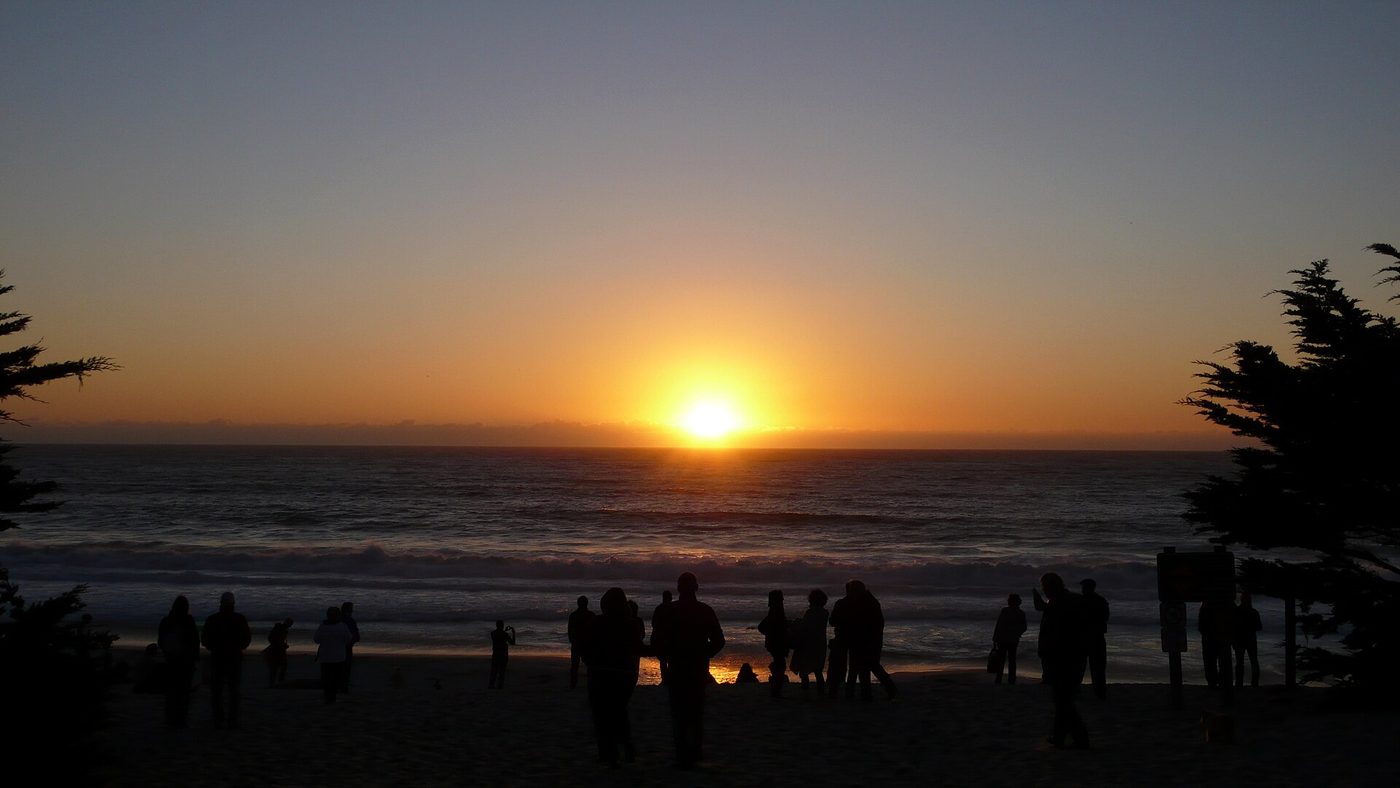Families watching the sun set over the water from the sand at Carmel Beach, with cypress branches framing the view